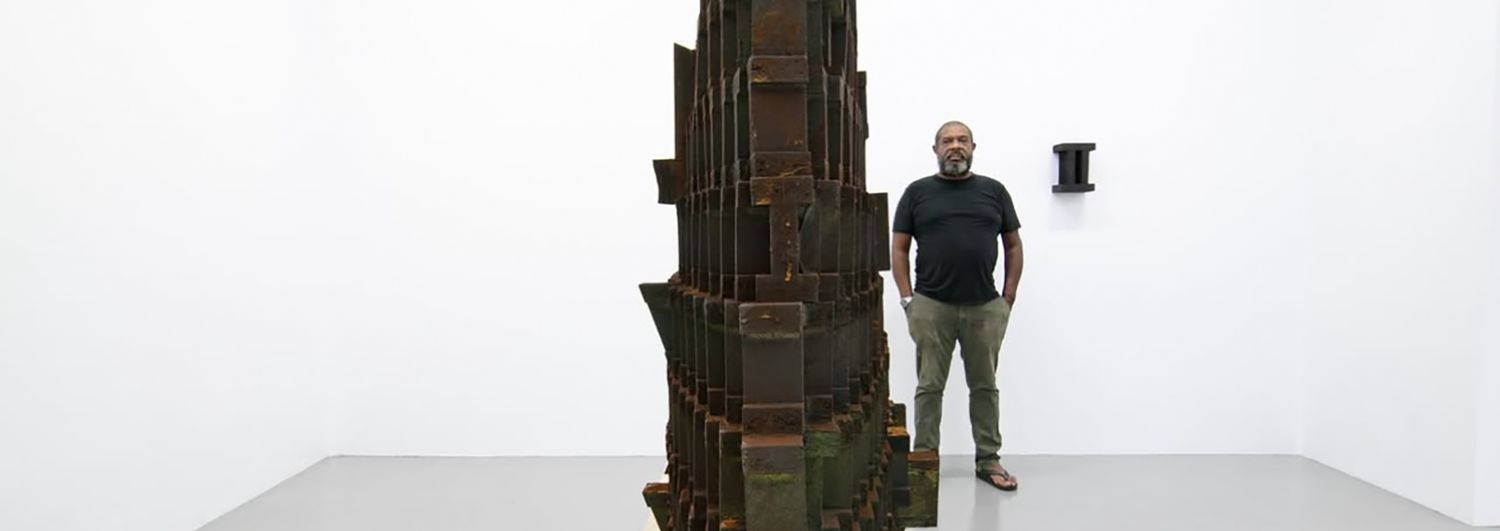 Thomas Sleet standing beside a tall charred and rusted wood sculpture in the gallery